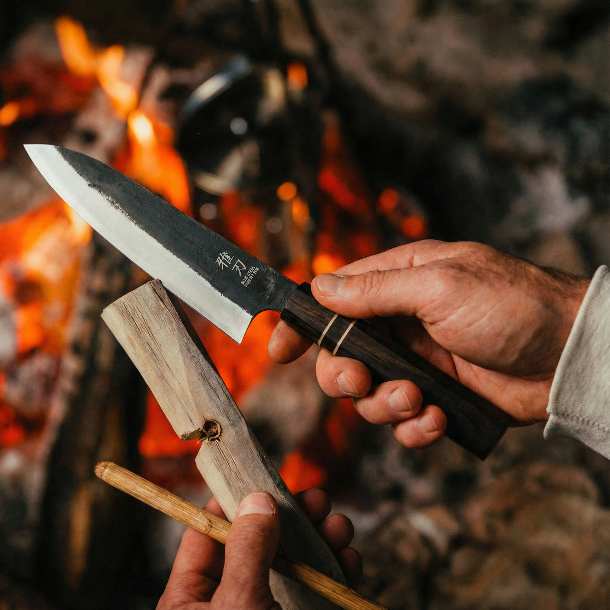 A person holding a Japanese Blue Steel petty knife while carving a piece of wood outdoors, with a glowing campfire blurred in the background.